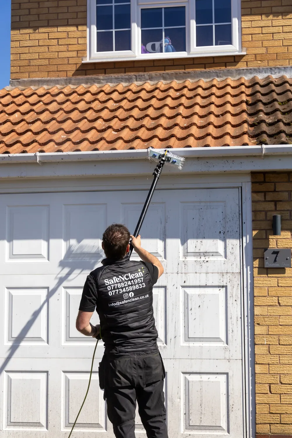 Operative in Safe'N'Clean uniform cleaning a window above a garage with an extended pole